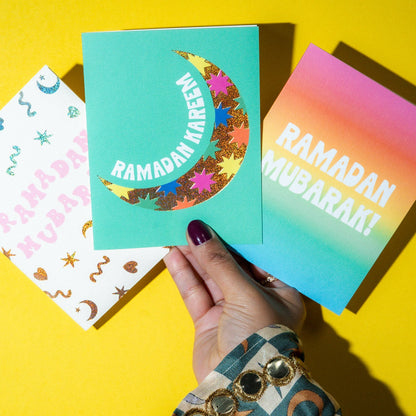 Three colorful Ramadan greeting cards held by a hand on a yellow background